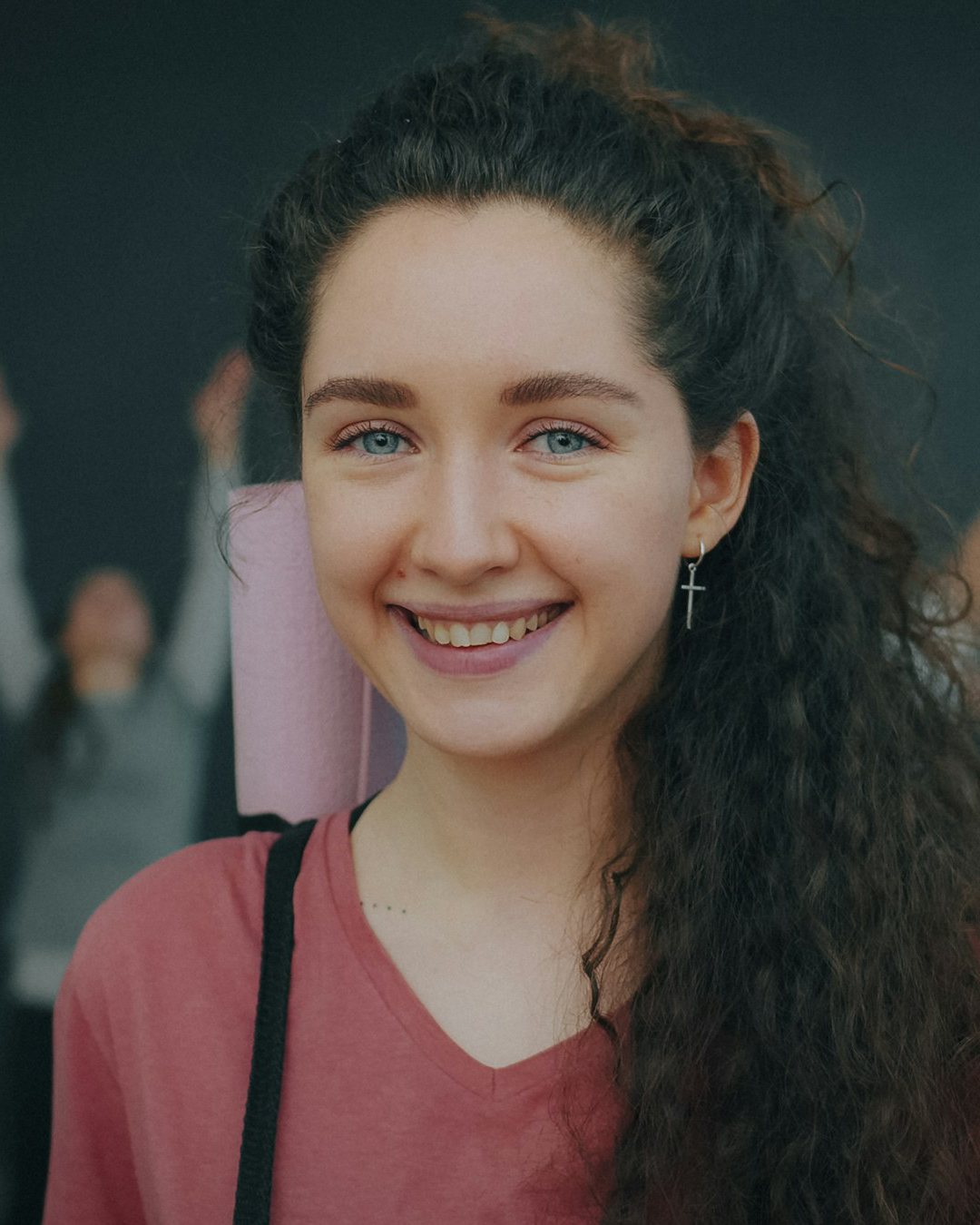 Nina, long curly hair and a red top, smiling with her mat in a class