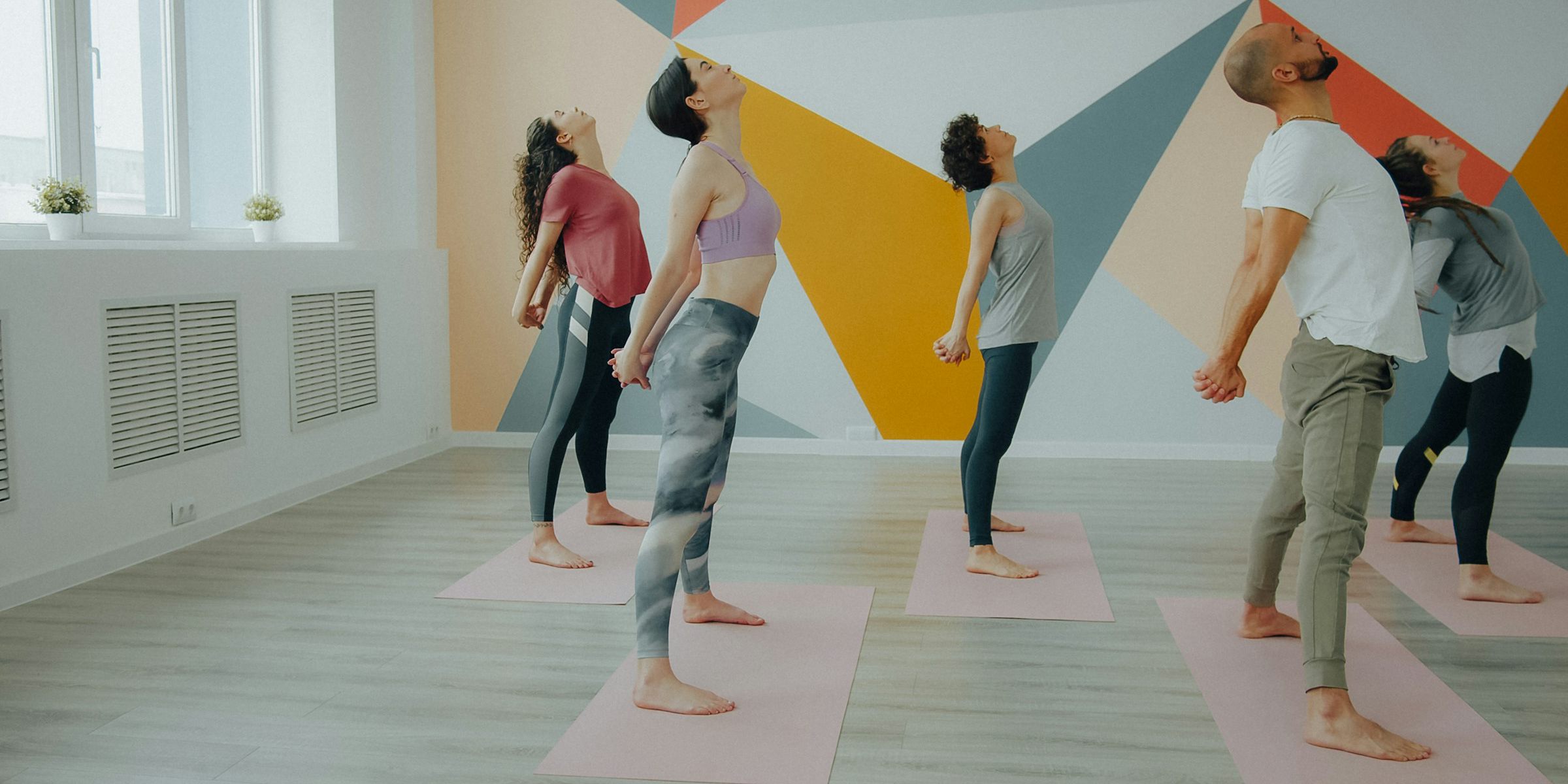 A class leaning back with arms behind them in front of a coral, orange and slate mural, pink mats on a wooden floor