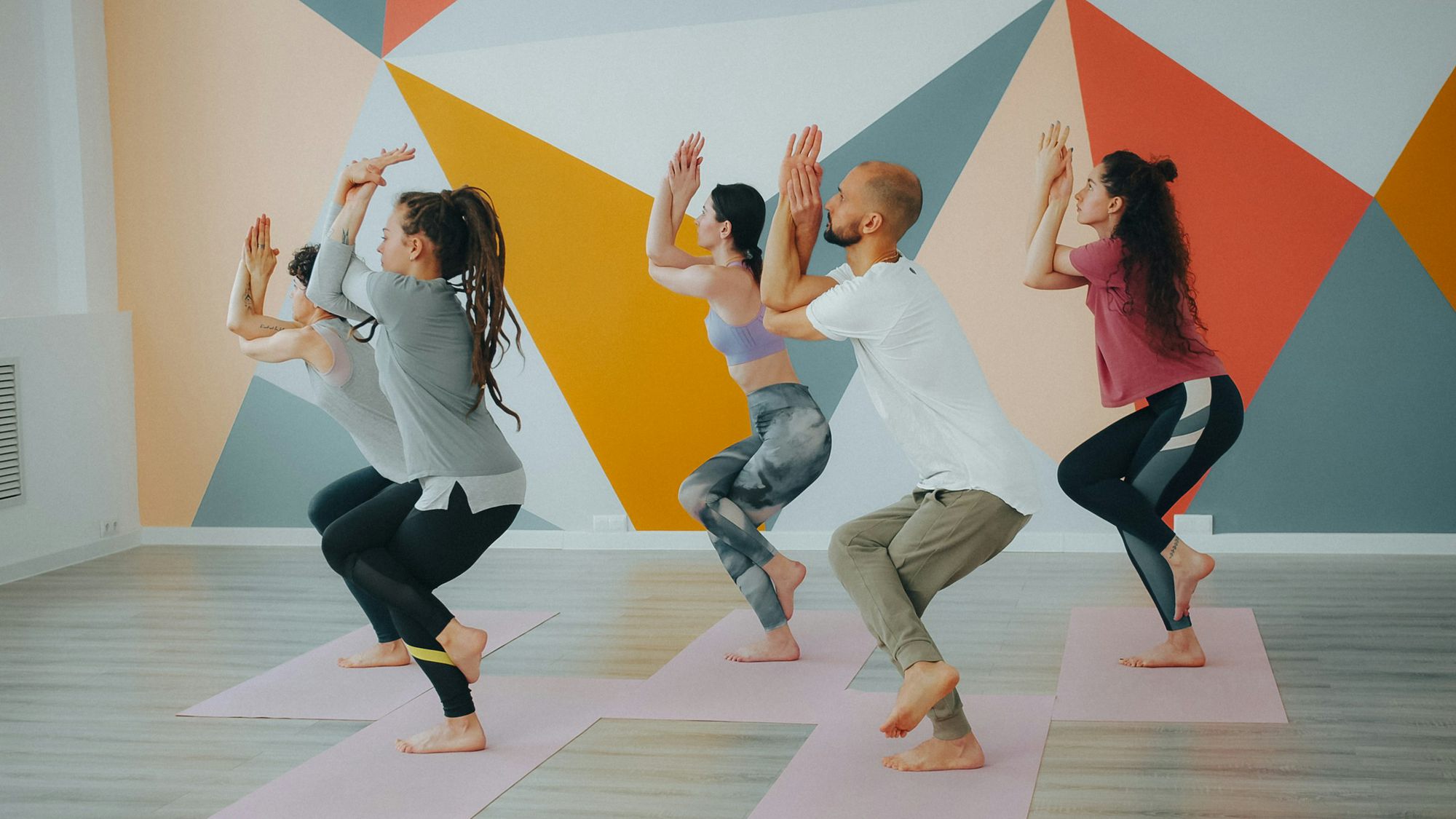 A class holding a chair pose in front of the coral and orange mural