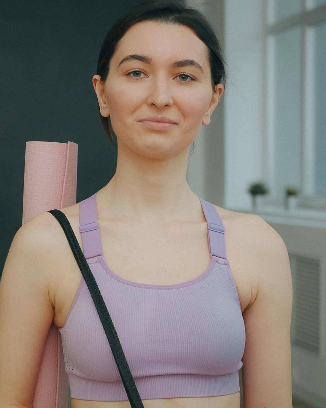 Mara, dark hair pulled back, in a lilac top with her mat on her shoulder
