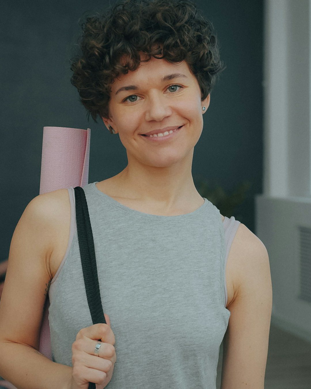 Ruth, short curly hair and a grey vest, smiling with her rolled mat on her shoulder
