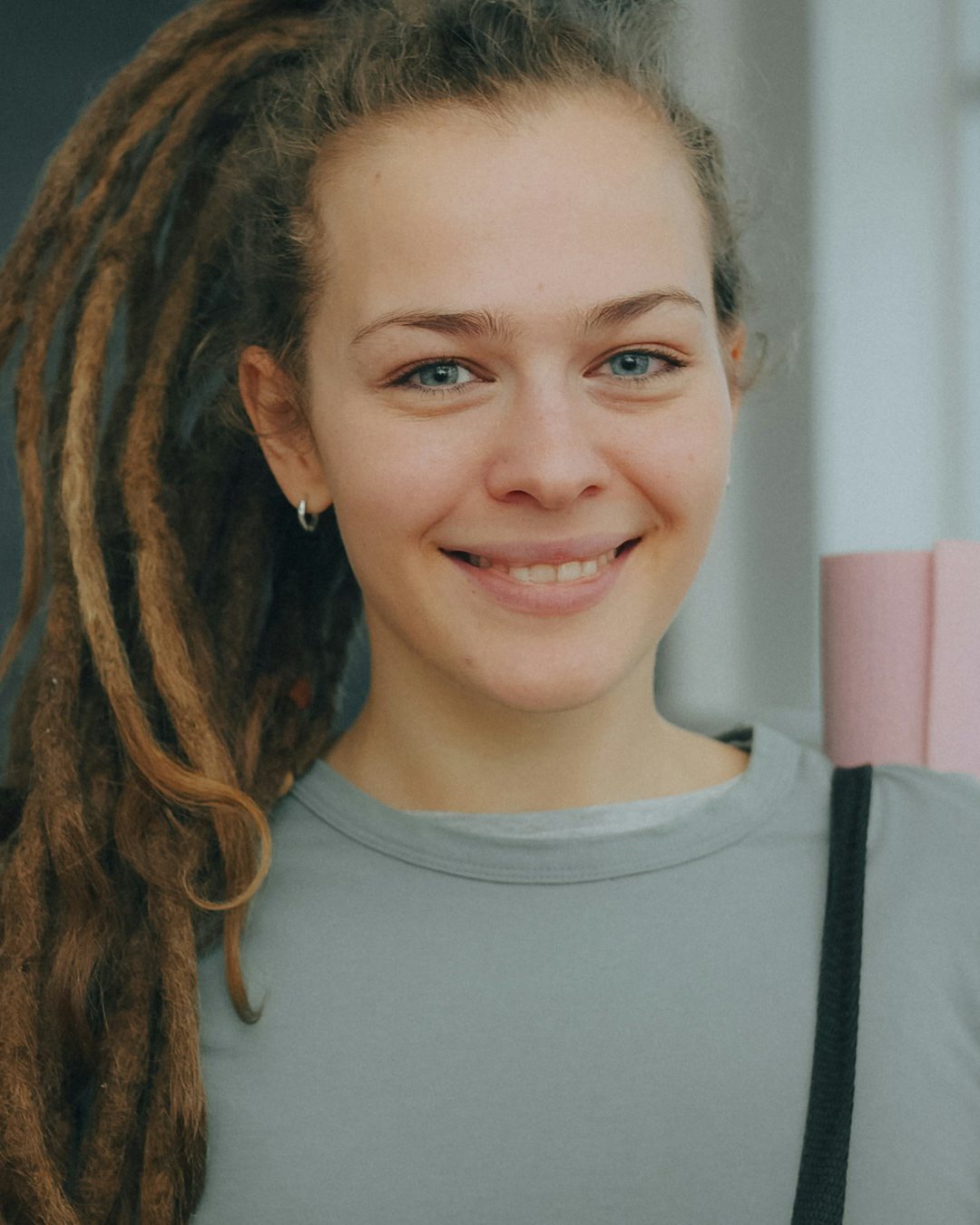 Tess, long dreadlocks and a grey top, smiling with her mat strap over her shoulder