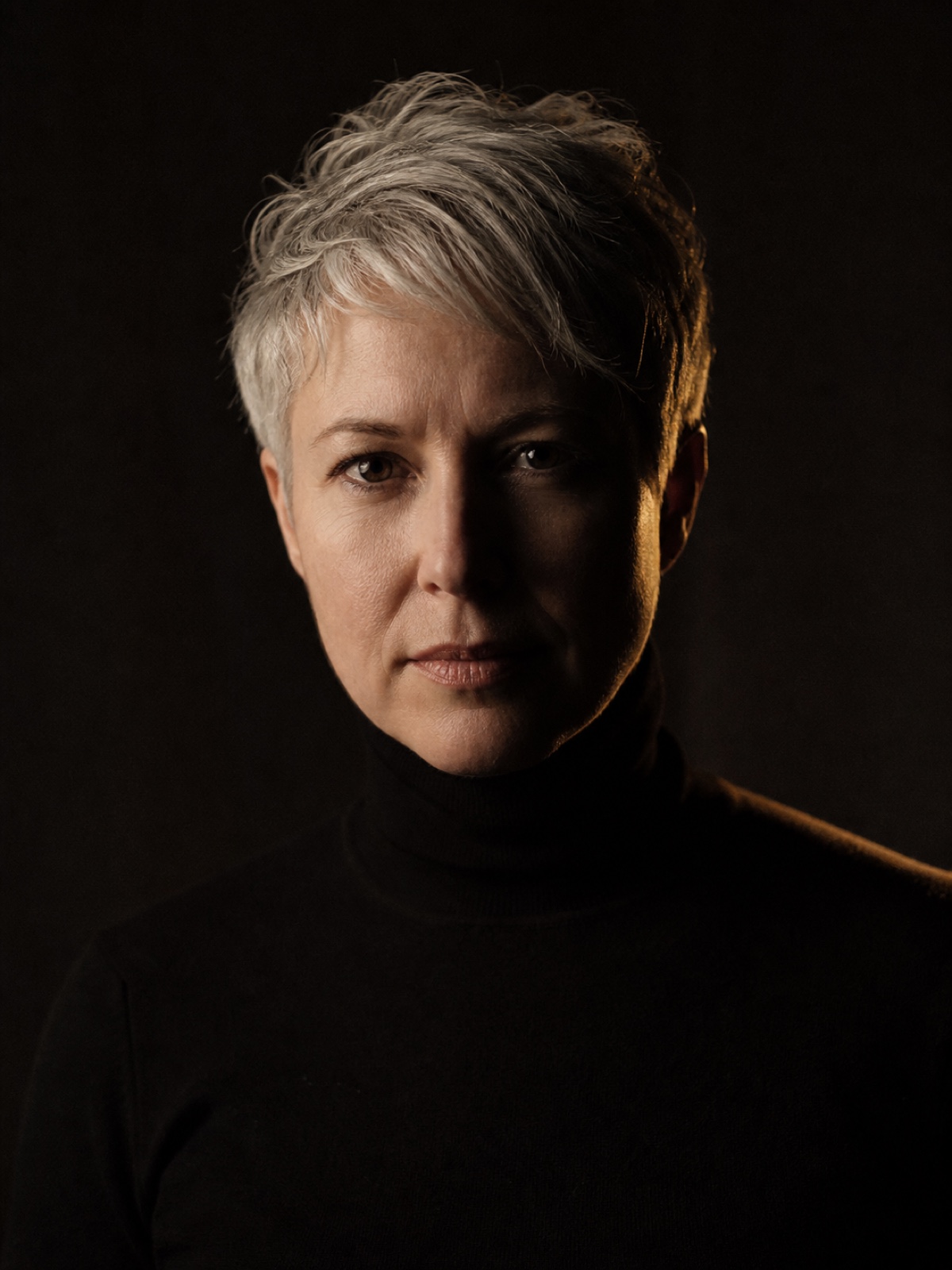 Editorial portrait of a woman in her forties with cropped silver hair and a black turtleneck, lit from one side against a deep charcoal backdrop