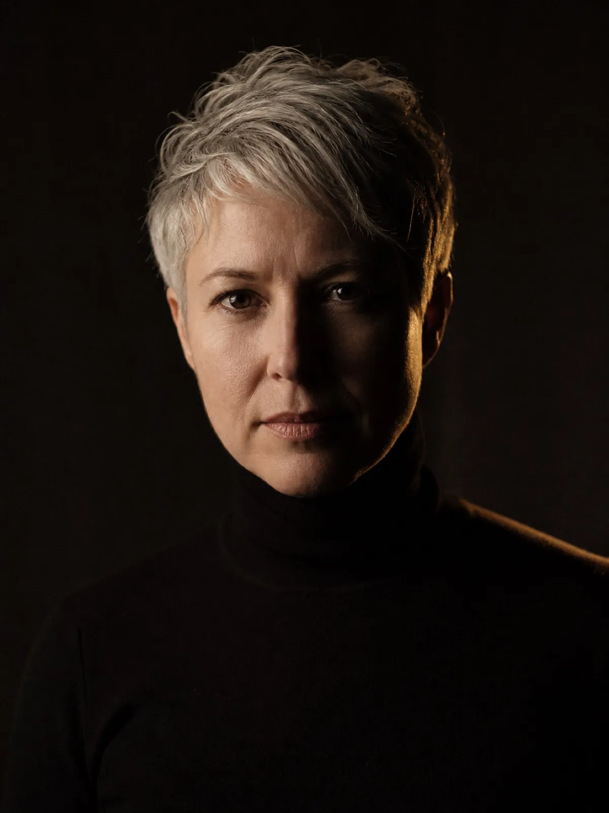 Editorial portrait of a woman in her forties with cropped silver hair and a black turtleneck, lit from one side against a deep charcoal backdrop