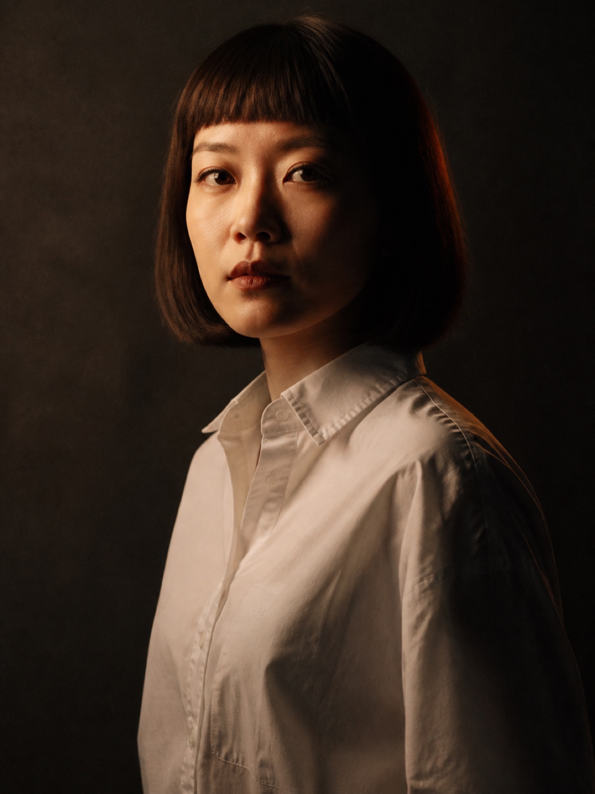 Editorial portrait of an East Asian woman in her thirties with a blunt bob and a white shirt, three-quarter turn, soft key light on a charcoal backdrop