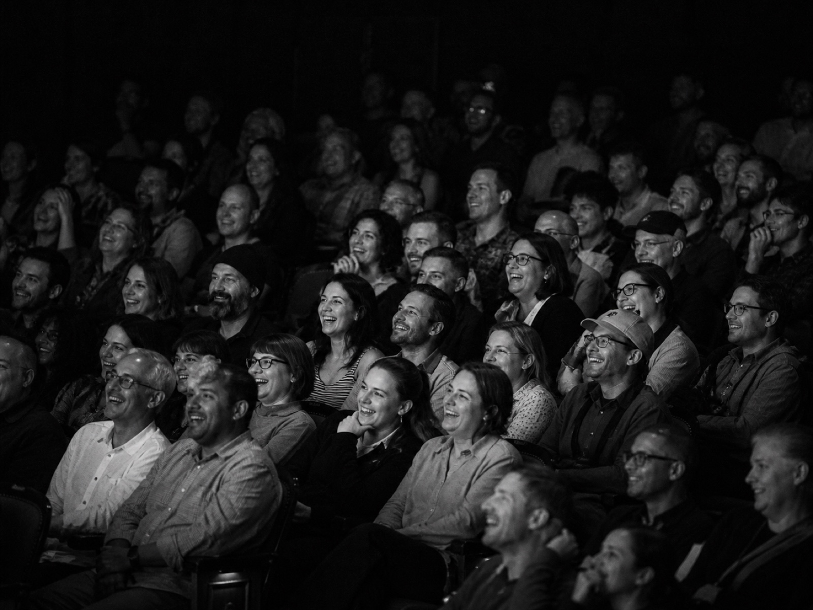 Black and white photograph of the audience mid-laugh, rows of faces lit only by stage spill