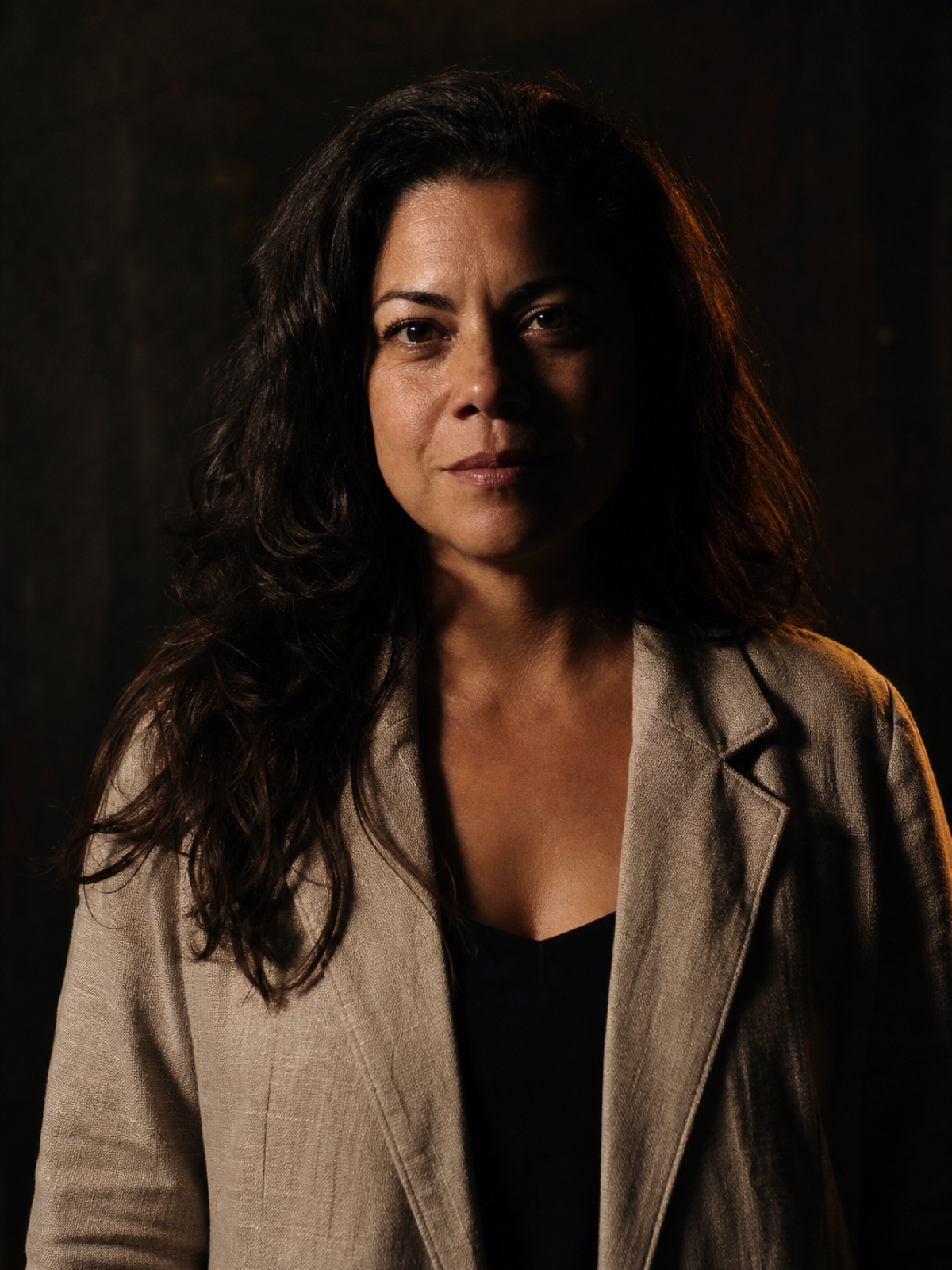 Editorial portrait of a Latina woman in her forties with dark wavy hair and a linen jacket, half-smile, warm rim light on a charcoal backdrop