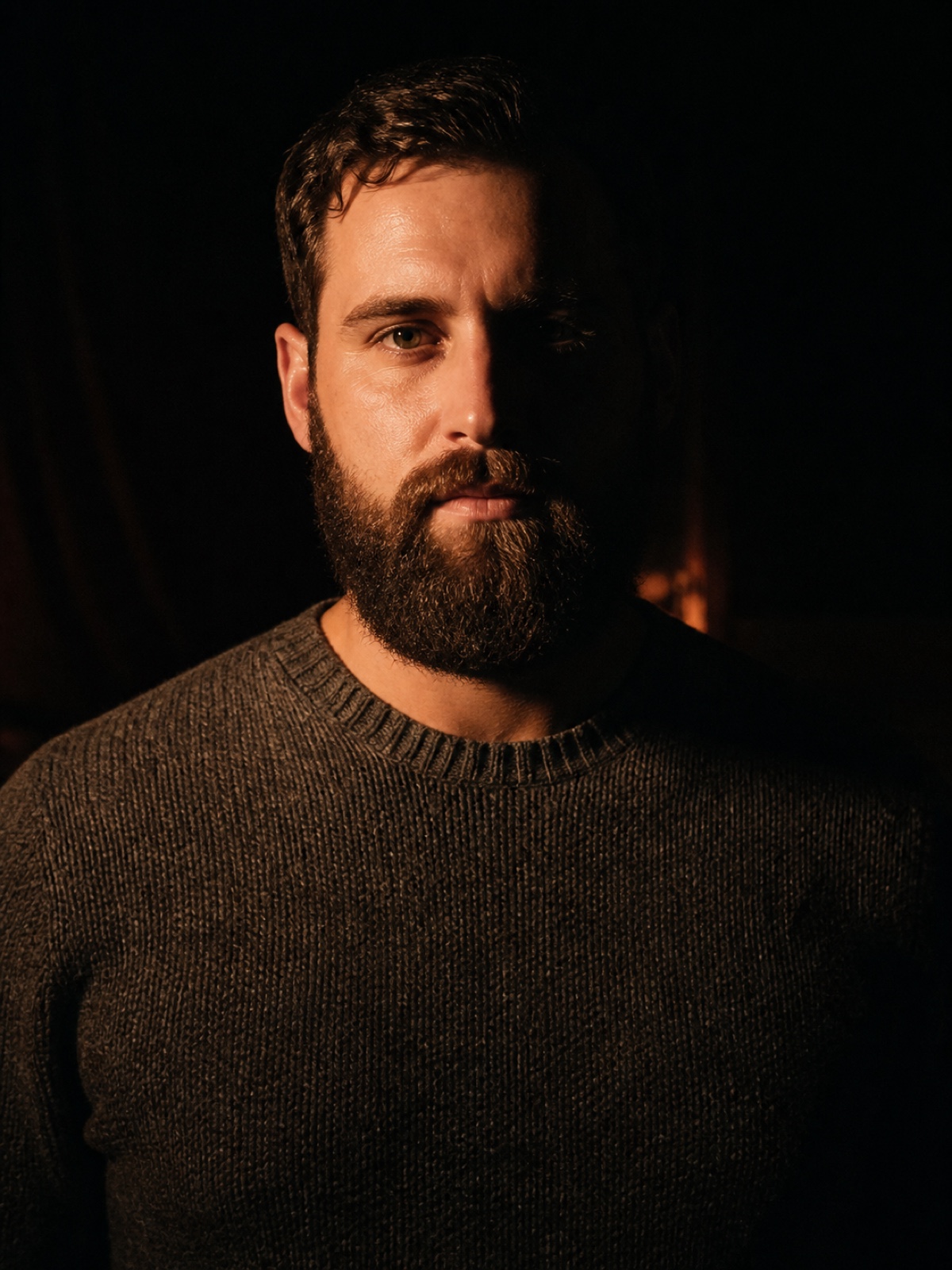 Editorial portrait of a bearded man in his thirties in a heavy knit sweater, half his face in shadow, deep charcoal backdrop