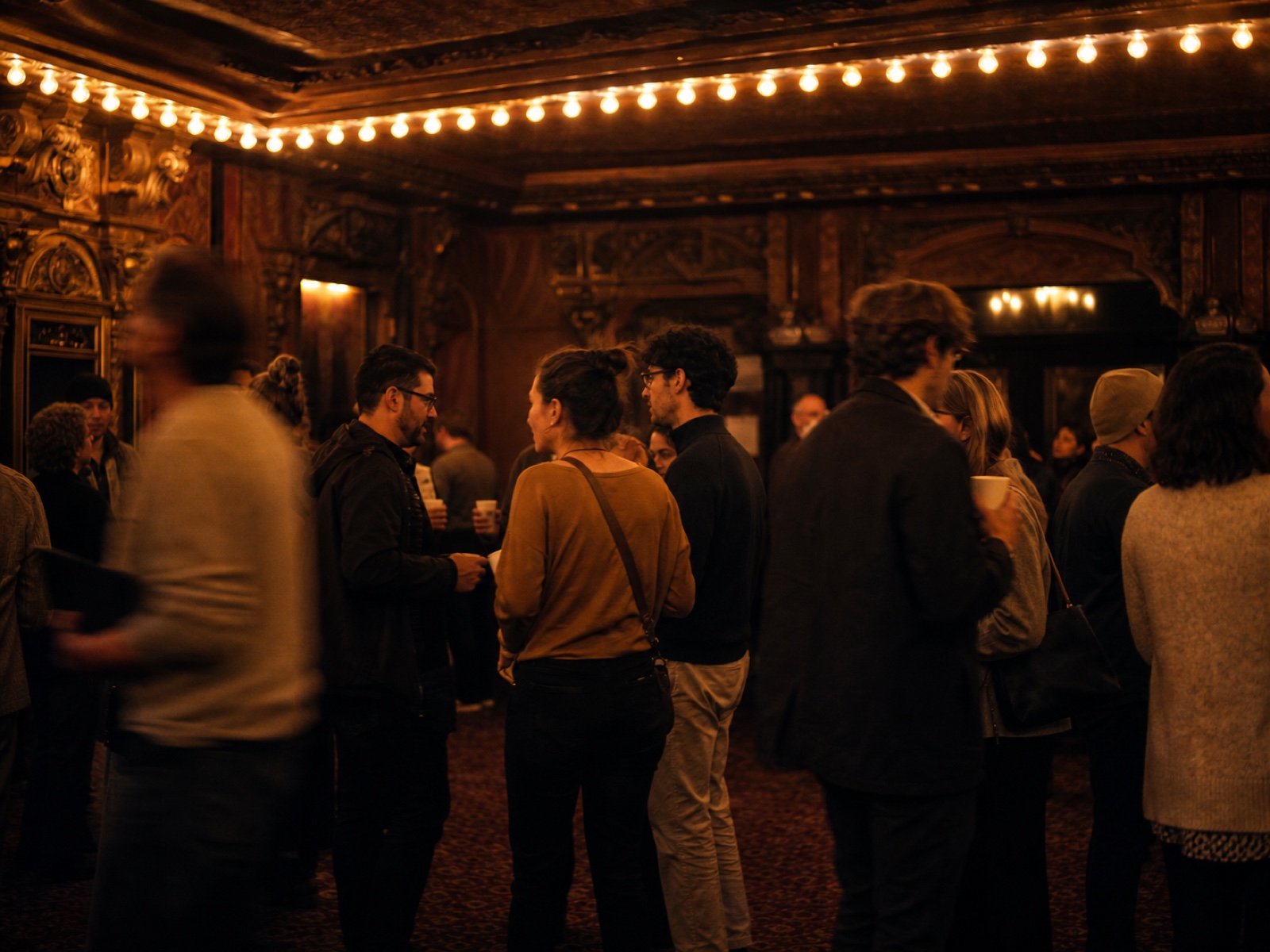 The theatre lobby between talks: attendees standing in clusters under a marquee bulb sign, warm tungsten light, coffee cups, motion blur in the crowd