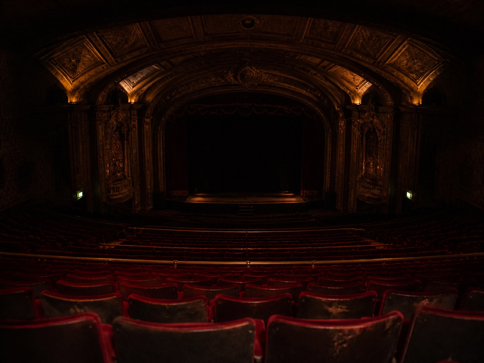 The empty auditorium from the back of the balcony: rows of red velvet seats curving away, house lights low, exit signs glowing