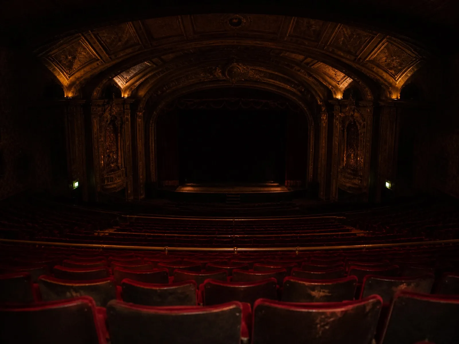 The empty auditorium from the back of the balcony: rows of red velvet seats curving away, house lights low, exit signs glowing