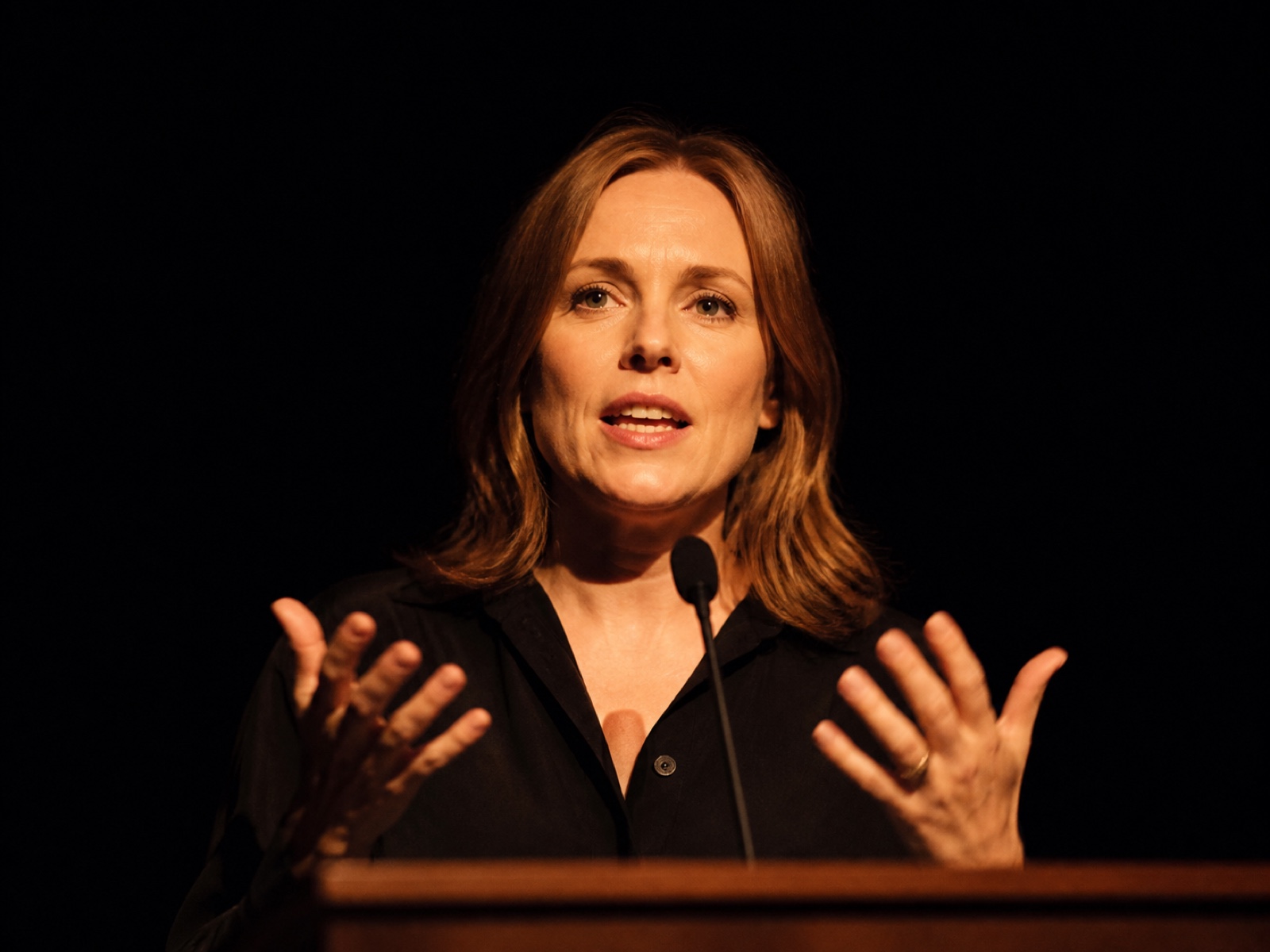 Close crop of a speaker mid-sentence at the lectern, hands raised, lit hard from one side against a black stage, shallow depth of field