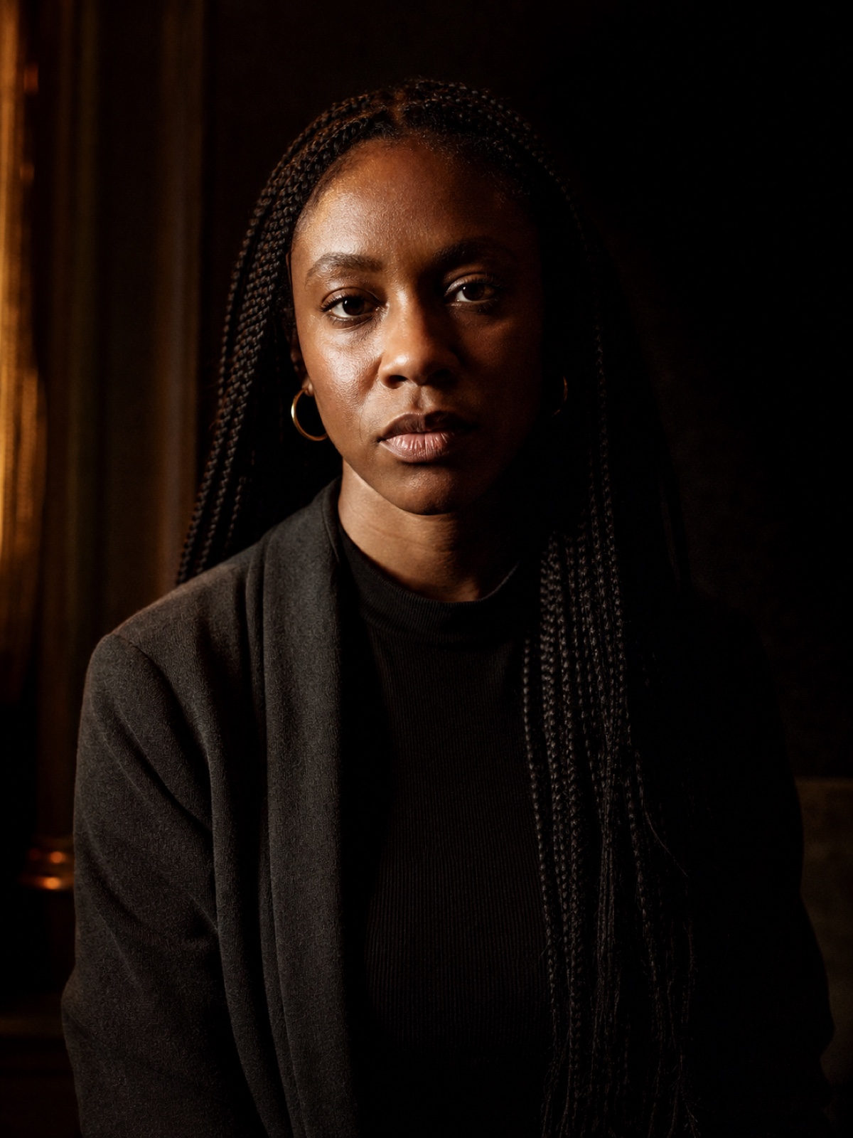 Editorial portrait of a Black woman in her thirties with long braids and gold hoop earrings, direct gaze, hard side light on a charcoal backdrop