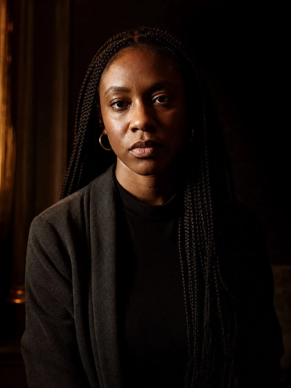 Editorial portrait of a Black woman in her thirties with long braids and gold hoop earrings, direct gaze, hard side light on a charcoal backdrop