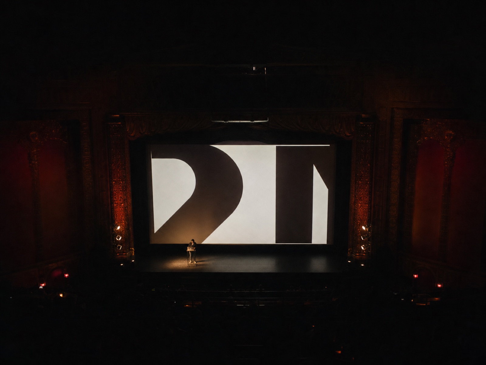 Wide shot of the stage from the balcony: a single speaker dwarfed by a huge projected slide of typography