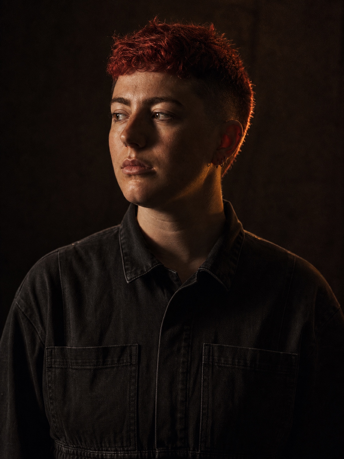 Editorial portrait of a non-binary person in their twenties with dyed hair and a boiler suit, looking off-camera, hard side light, charcoal backdrop