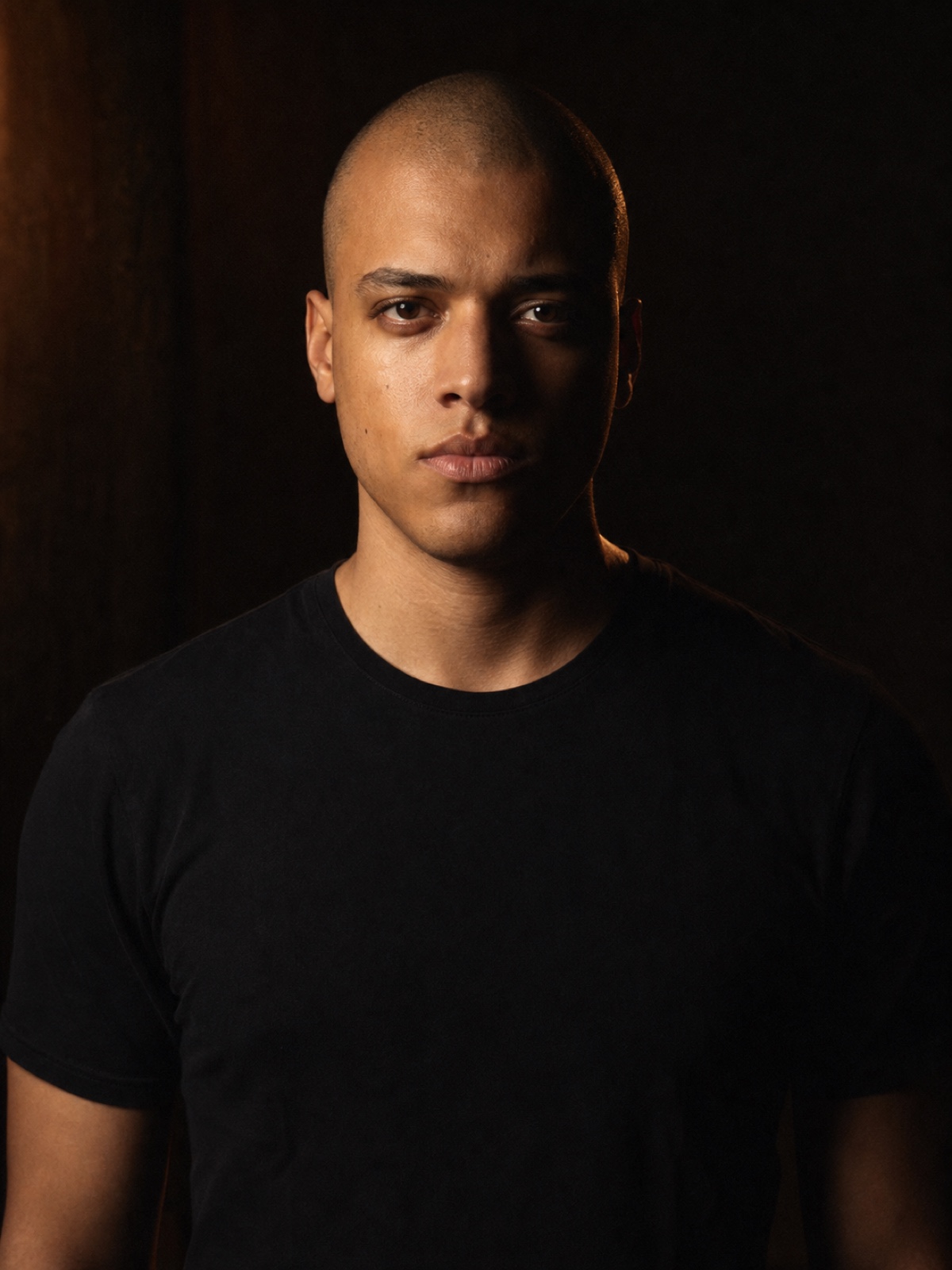 Editorial portrait of a man in his twenties with a shaved head and a plain black t-shirt, shoulders square to camera, charcoal backdrop