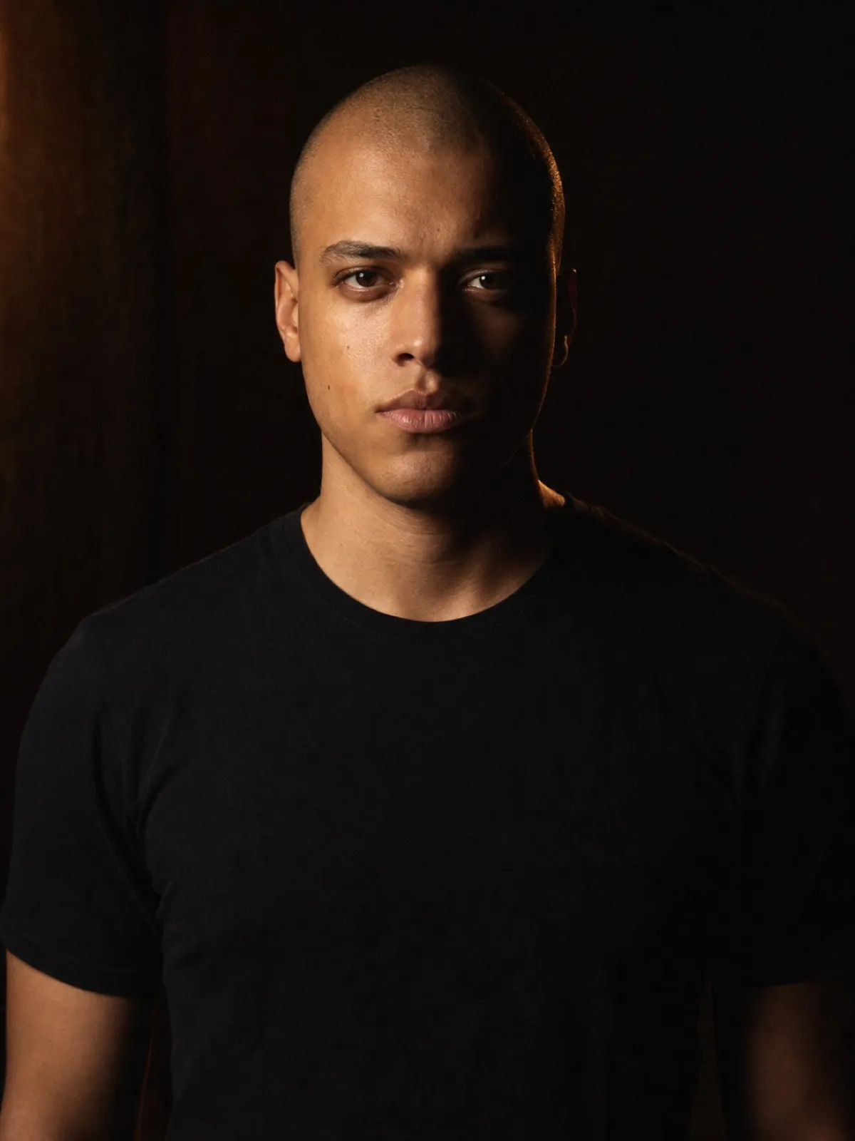 Editorial portrait of a man in his twenties with a shaved head and a plain black t-shirt, shoulders square to camera, charcoal backdrop