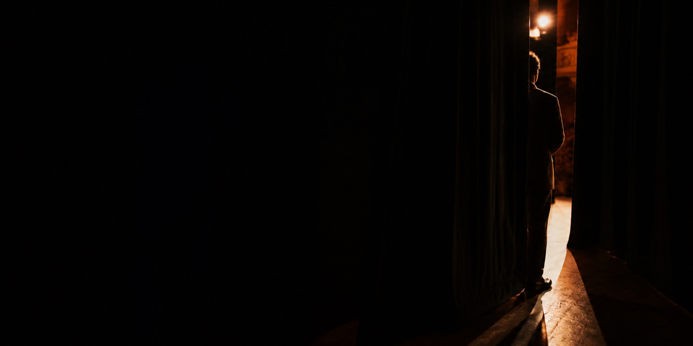 A speaker waiting in the wings, lit only by the stage spill, watching the previous talk from behind the curtain