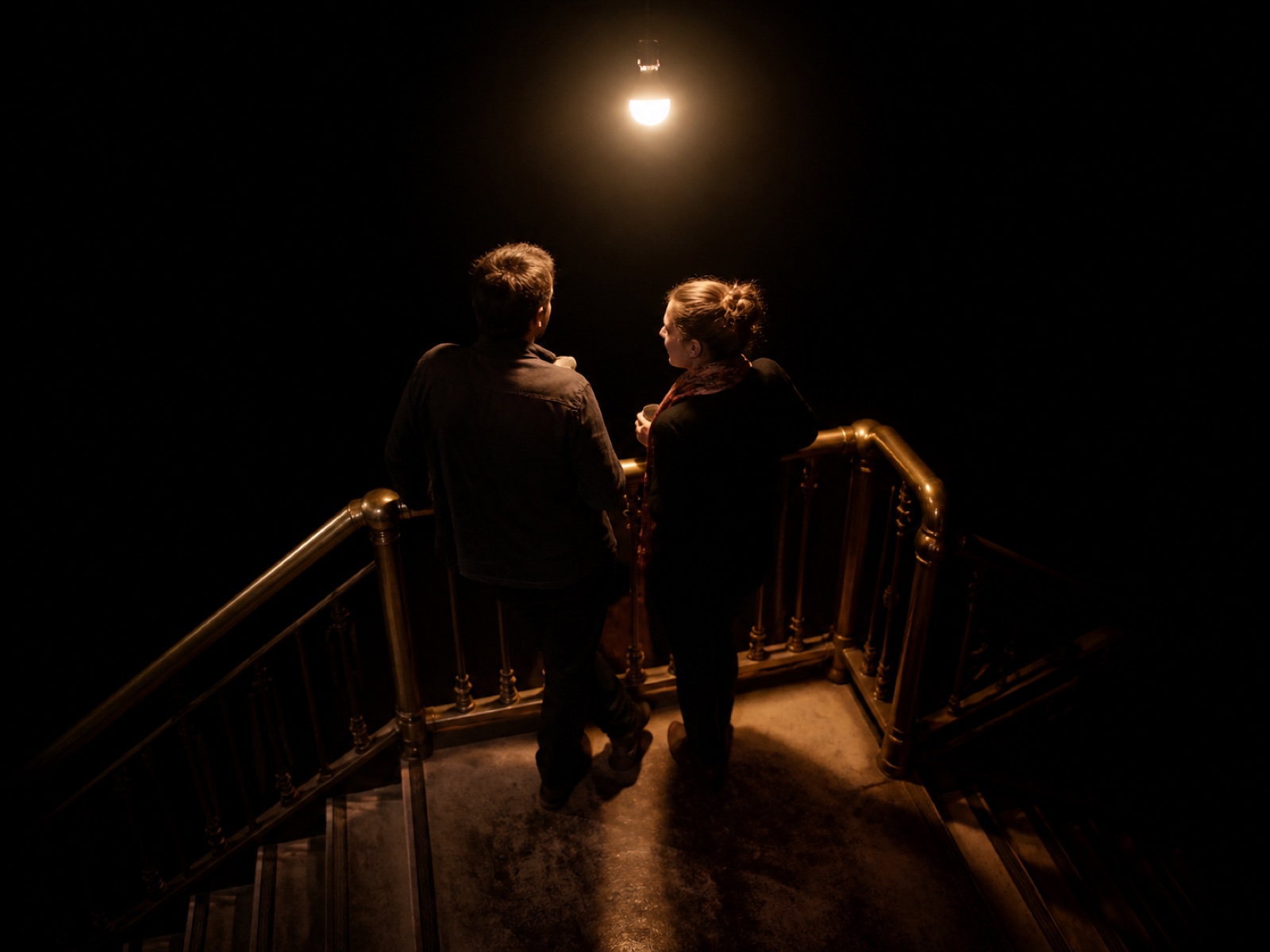 Two attendees talking closely in a stairwell, warm bulb light overhead, everything else dark