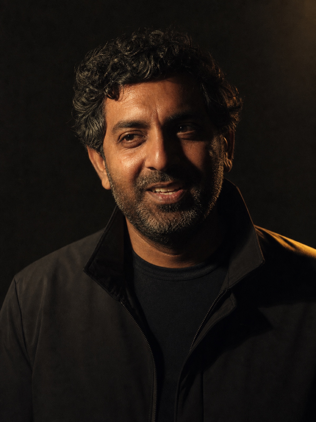 Editorial portrait of a South Asian man in his forties in a dark jacket, laughing slightly, hard directional light on a deep charcoal backdrop