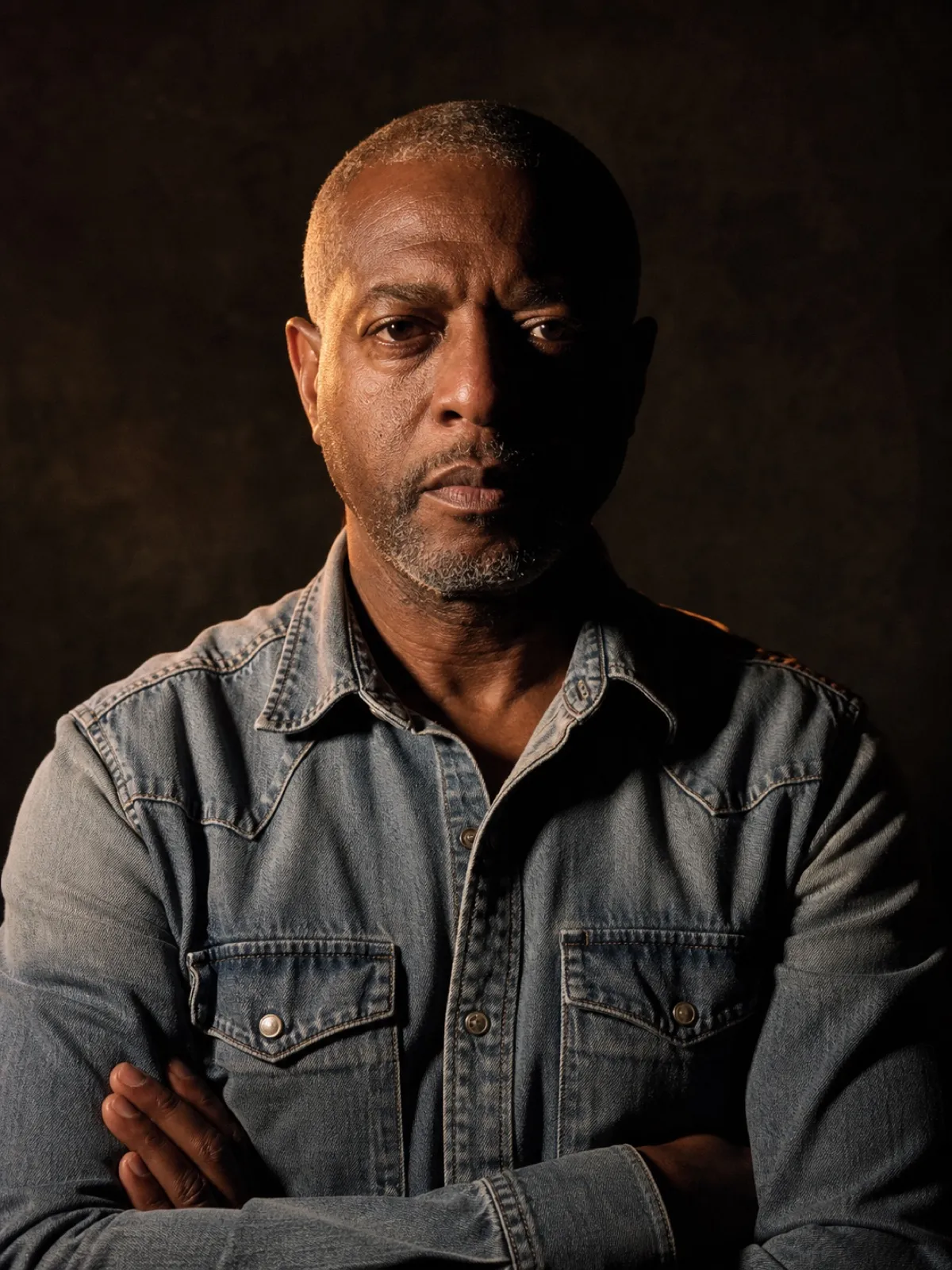 Editorial portrait of a Black man in his fifties with close-cropped grey hair and a denim shirt, arms folded, warm rim light on a charcoal backdrop