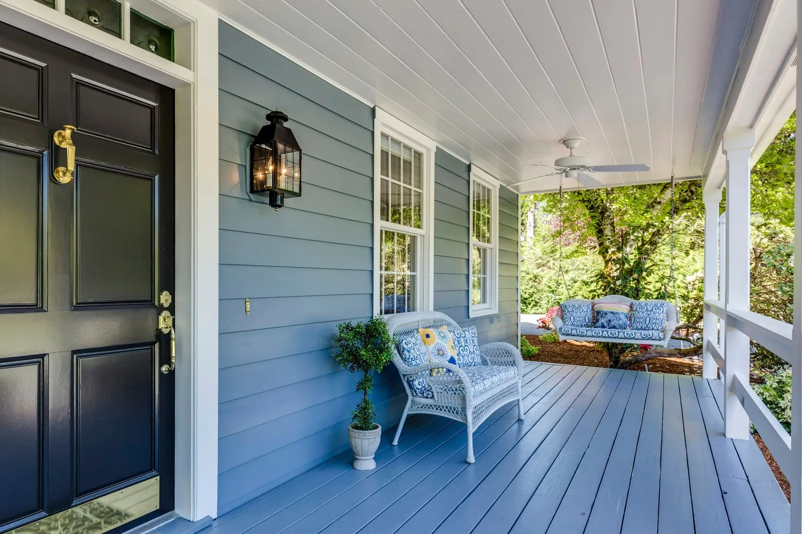 A blue-gray porch with a wicker chair and a black front door