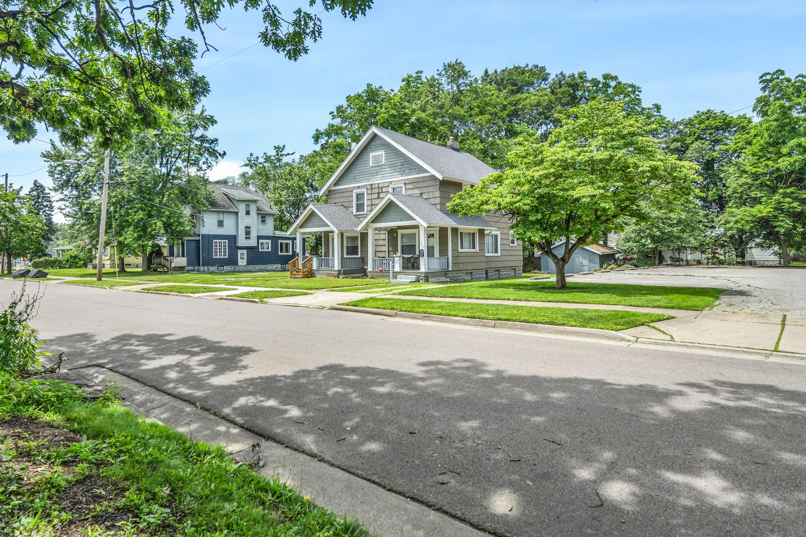 A gray shingled house with a front porch on a wide, leafy street
